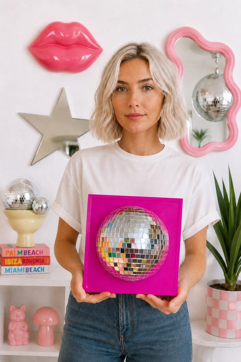 May include: A square, bright pink wall-mounted frame featuring a centered, spherical disco ball made of small, reflective silver mirror tiles. The disco ball is securely attached to the flat, solid pink square base, creating a dimensional effect.