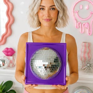 May include: A square purple wall-mounted frame featuring a centered, hemispherical silver disco ball made of small reflective mirror tiles. The frame has a smooth, solid purple border surrounding the circular, faceted mirrored surface.