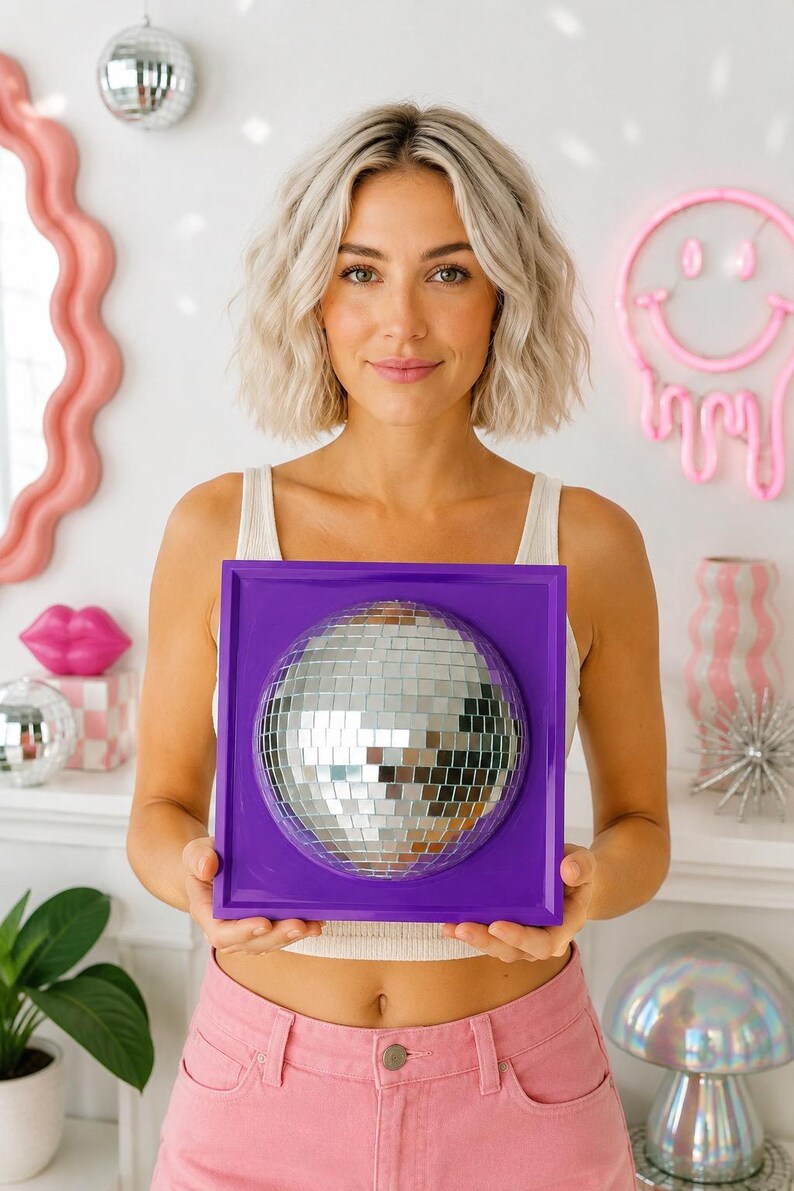 May include: A square purple wall-mounted frame featuring a centered, hemispherical silver disco ball made of small reflective mirror tiles. The frame has a smooth, solid purple border surrounding the circular, faceted mirrored surface.