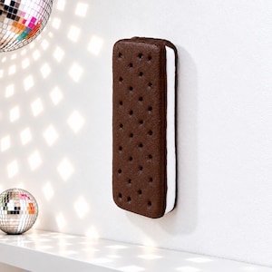 May include: A rectangular ice cream sandwich wall decor piece featuring two dark brown chocolate cookie wafers with a dotted texture and a thick layer of white cream filling in the center, mounted vertically on a plain white wall.