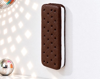 May include: A rectangular ice cream sandwich wall decor piece featuring two dark brown chocolate cookie wafers with a dotted texture and a thick layer of white cream filling in the center, mounted vertically on a plain white wall.