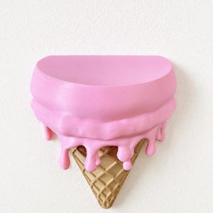 May include: A wall-mounted shelf shaped like an upside-down ice cream cone. The top section is a smooth, semi-circular pink surface with realistic pink drips cascading down over a textured, tan-colored waffle cone base that tapers to a point.