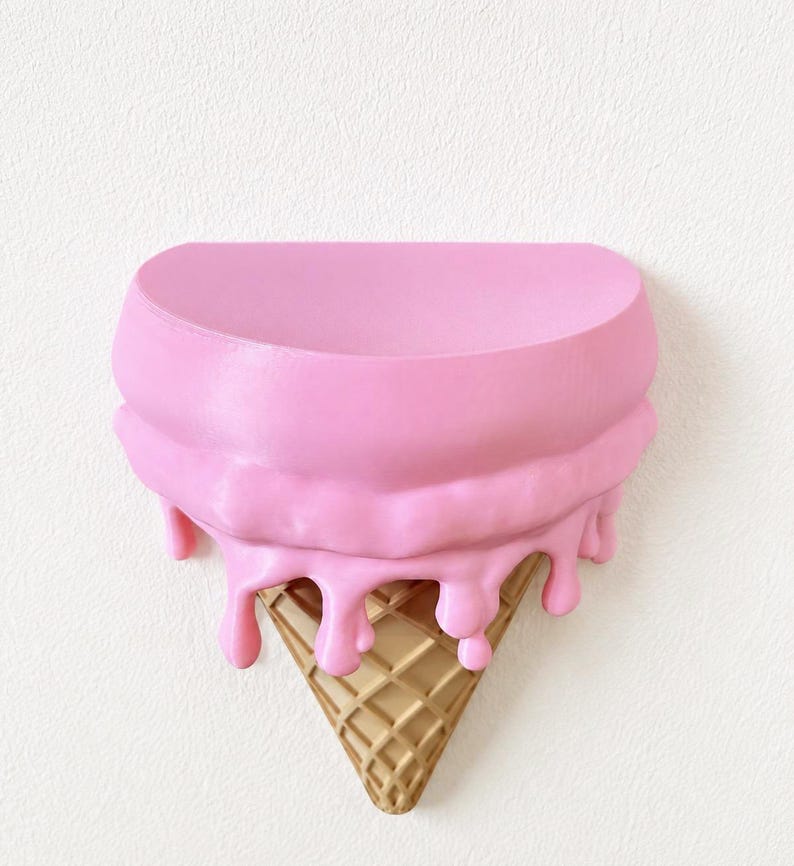 May include: A wall-mounted shelf shaped like an upside-down ice cream cone. The top section is a smooth, semi-circular pink surface with realistic pink drips cascading down over a textured, tan-colored waffle cone base that tapers to a point.