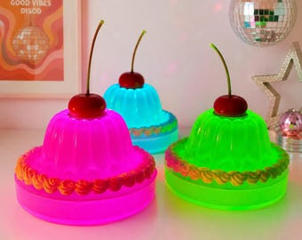 May include: Three illuminated jelly-shaped lamps in bright pink, blue, and green. Each lamp features a fluted base, a decorative piped border around the bottom, and a single red cherry topper with a long stem. The lamps emit a vibrant glow.