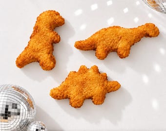 May include: Three golden-brown, breaded dinosaur-shaped chicken nuggets arranged on a white surface, accompanied by two reflective, mirrored disco balls of different sizes positioned in the corners of the frame.
