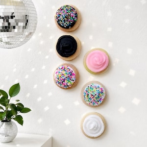 May include: Six round cookies arranged vertically on a wall. The top cookie has black frosting with multicolored sprinkles, followed by a plain black frosted cookie, a pink frosted cookie, a cookie with multicolored sprinkles, another with sprinkles, and a white frosted cookie.