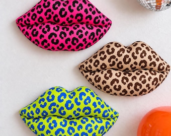 May include: Three plush lip-shaped pillows featuring animal print patterns. One pillow is bright pink with black spots, one is tan with brown spots, and one is lime green with blue spots. Each pillow has a distinct, soft, rounded lip shape.