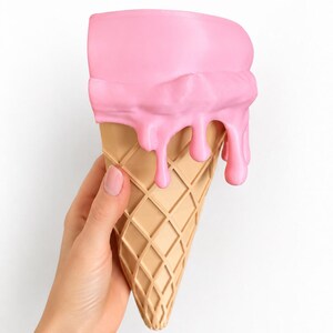 May include: A decorative object shaped like an ice cream cone with a tan, waffle-textured base and a smooth, bright pink scoop on top. The pink portion features realistic, thick drips cascading down the sides of the cone, creating a textured effect.