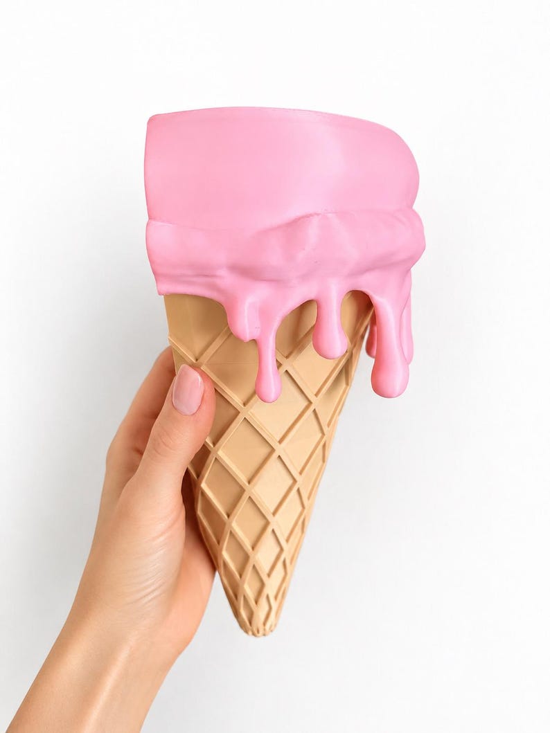 May include: A decorative object shaped like an ice cream cone with a tan, waffle-textured base and a smooth, bright pink scoop on top. The pink portion features realistic, thick drips cascading down the sides of the cone, creating a textured effect.