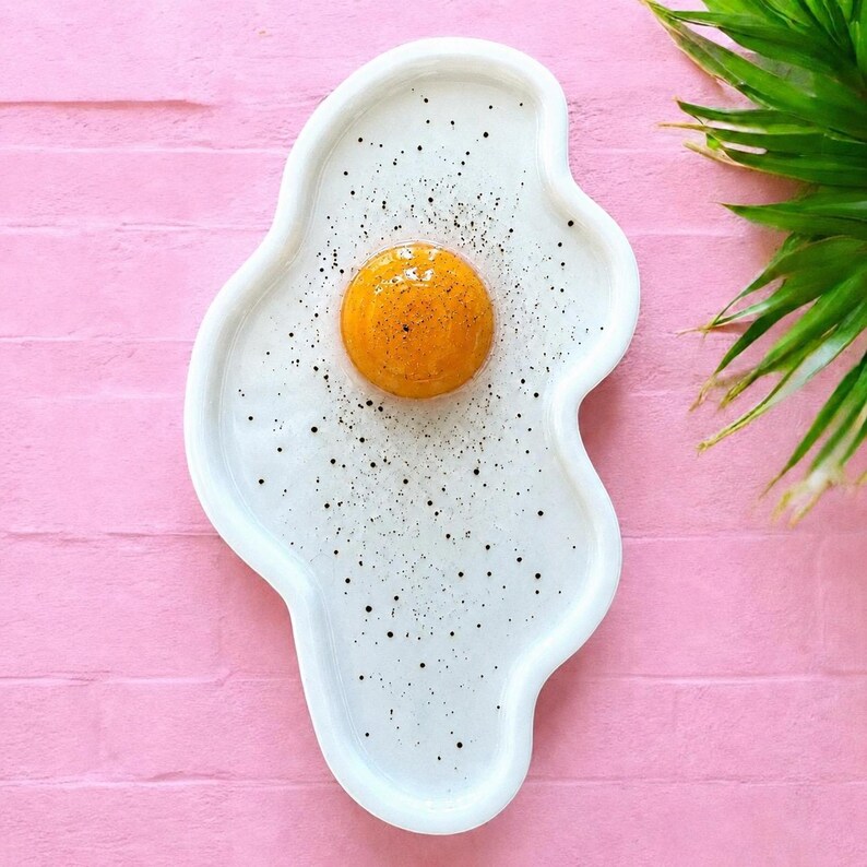 Puede incluir: Un plato de servicio de cer&aacute;mica blanca con forma de huevo frito y contorno irregular y org&aacute;nico. El centro presenta una yema naranja brillante, elevada y lustrosa, mientras que la clara est&aacute; salpicada de peque&ntilde;os puntos negros.