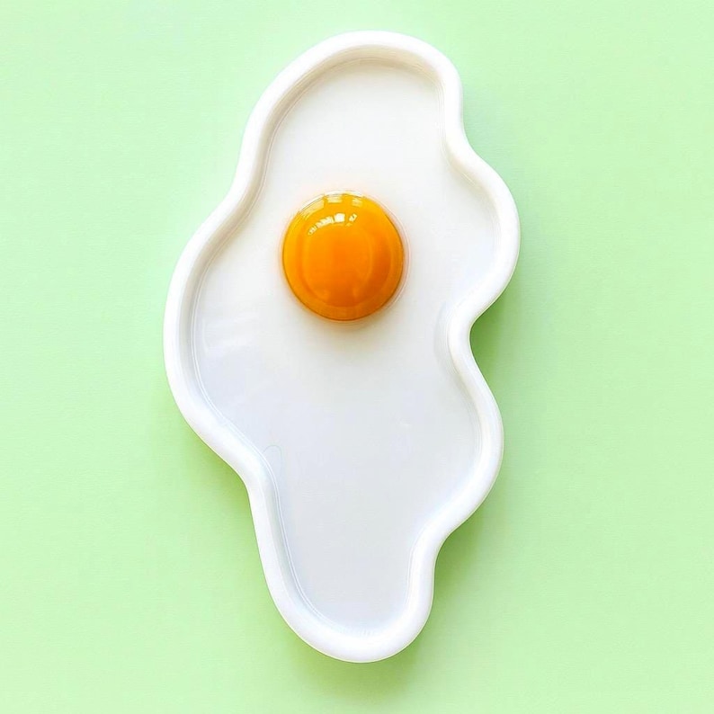 Puede incluir: Un plato de cer&aacute;mica blanca con forma de huevo frito y contorno irregular y org&aacute;nico. El centro presenta un elemento circular elevado, brillante y de color amarillo anaranjado intenso que destaca sobre la superficie lisa y blanca.