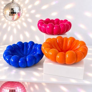 May include: Three glossy, rounded, scalloped bowls in vibrant solid colors: one bright pink, one deep blue, and one vivid orange. Each bowl features a continuous, bubble-like rim and a smooth, reflective surface, arranged in a cluster.