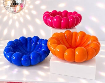 May include: Three glossy, rounded, scalloped bowls in vibrant solid colors: one bright pink, one deep blue, and one vivid orange. Each bowl features a continuous, bubble-like rim and a smooth, reflective surface, arranged in a cluster.