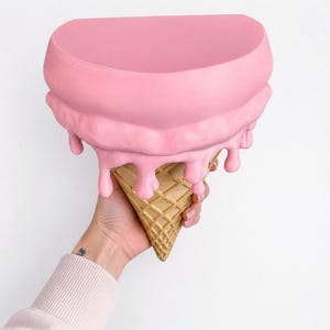 May include: A semi-circular wall shelf shaped like an ice cream cone. The top surface is flat and pink, with sculpted pink drips cascading down the sides onto a tan, textured waffle cone base that tapers to a point at the bottom.