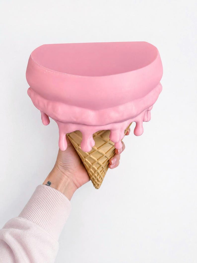 May include: A semi-circular wall shelf shaped like an ice cream cone. The top surface is flat and pink, with sculpted pink drips cascading down the sides onto a tan, textured waffle cone base that tapers to a point at the bottom.