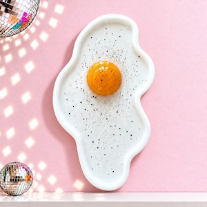 Puede incluir: Una bandeja de cer&aacute;mica con forma de huevo frito con un borde blanco irregular, una yema naranja brillante en relieve en el centro y peque&ntilde;as motas negras esparcidas por la superficie blanca que parecen pimienta.