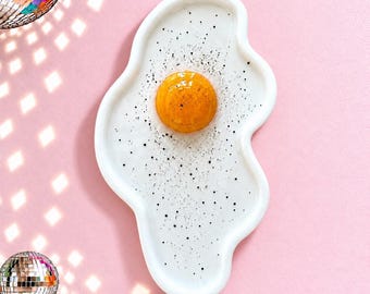 May include: A ceramic tray shaped like a fried egg with an irregular white border, a raised glossy orange yolk in the center, and small black speckles scattered across the white surface to resemble cracked black pepper.