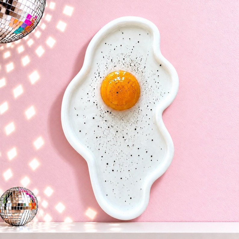 Puede incluir: Una bandeja de cer&aacute;mica con forma de huevo frito con un borde blanco irregular, una yema naranja brillante en relieve en el centro y peque&ntilde;as motas negras esparcidas por la superficie blanca que parecen pimienta.