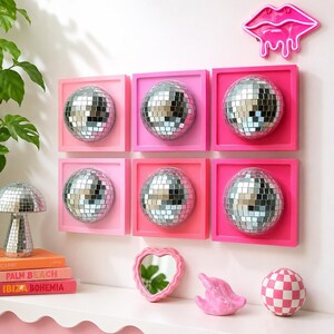 Pode incluir: Seis molduras de parede quadradas em v&aacute;rios tons de rosa, cada uma contendo uma esfera de globo de discoteca prateada. Um letreiro de n&eacute;on de l&aacute;bios rosa, um candeeiro de cogumelo de espelhos, um espelho em forma de cora&ccedil;&atilde;o, uma escultura rosa e uma bola xadrez rosa e branca.
