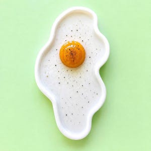 Puede incluir: Un plato de cer&aacute;mica blanco con forma de huevo frito irregular, con una yema central de color amarillo anaranjado brillante en relieve y peque&ntilde;as motas negras esparcidas por la superficie blanca como pimienta.