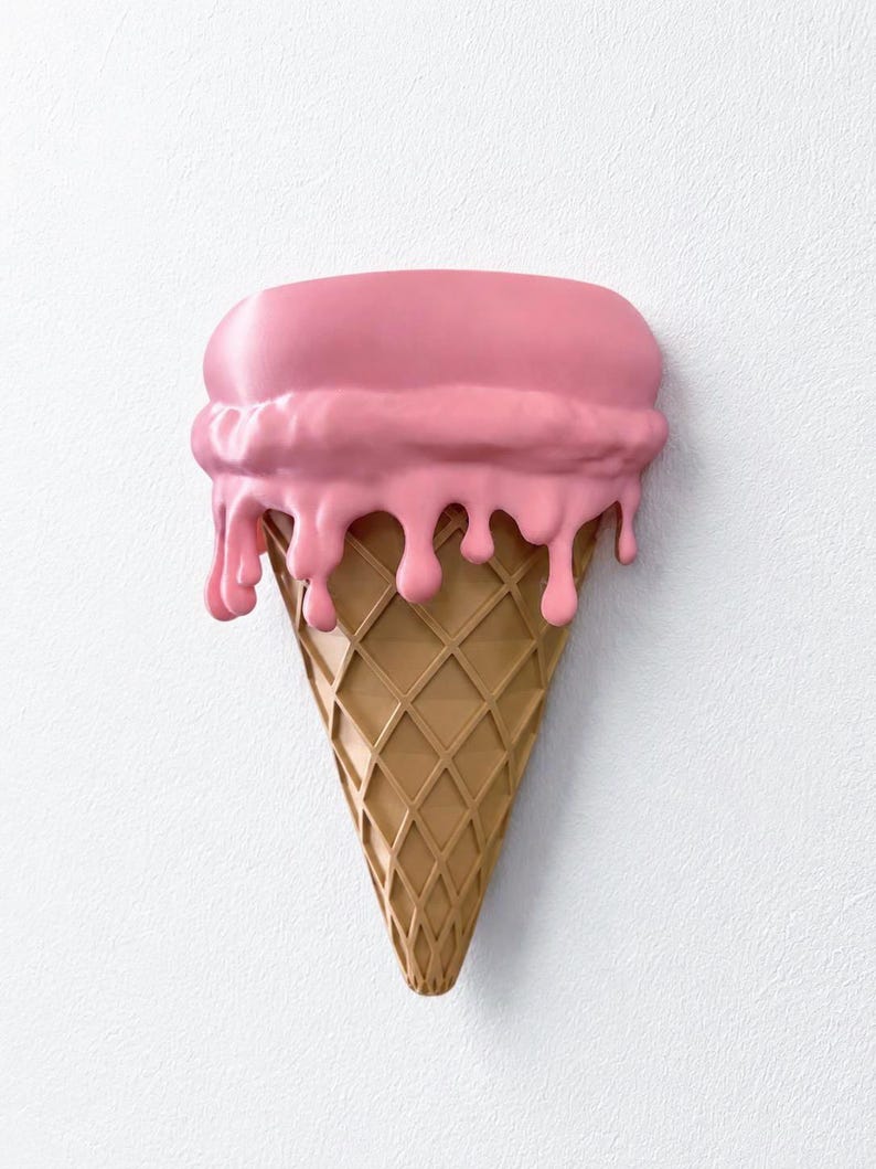 May include: A wall-mounted sculpture shaped like an ice cream cone with a tan, waffle-textured base and a large scoop of smooth, pink ice cream on top. The pink ice cream features realistic drips cascading down the sides of the cone.