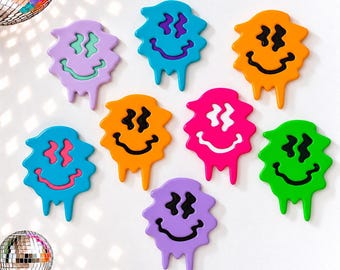 May include: Eight wall-mounted decorations shaped like melting smiley faces in various colors including purple, blue, orange, pink, and green. Each piece features wavy eyes and a curved mouth, with drips extending from the bottom edges.