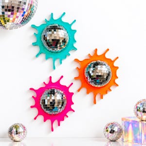 May include: Three wall-mounted disco balls featuring vibrant, paint-like drip frames in turquoise, orange, and hot pink. Each circular mirror ball is surrounded by a glossy, irregular splash-shaped border. Additional disco balls are placed below.