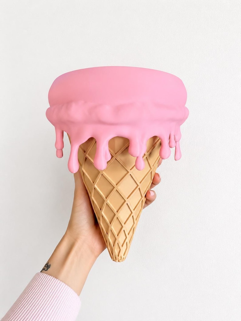 May include: A decorative object shaped like an ice cream cone with a tan, waffle-textured base and a smooth, matte pink top section featuring realistic drips cascading down the sides of the cone. The item has a rounded, flat top surface.