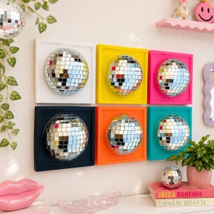 May include: Six square wall art pieces, each featuring a centered, reflective silver disco ball. The frames are colored white, yellow, pink, dark blue, orange, and teal. Each disco ball is composed of many small, square, mirrored glass tiles.