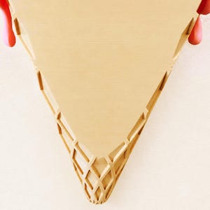 May include: A tan, triangular wall-mounted shelf shaped like an ice cream cone. The surface features a textured, waffle-like grid pattern at the pointed base and two keyhole slots at the top for mounting. Pink, rounded accents protrude from the sides.