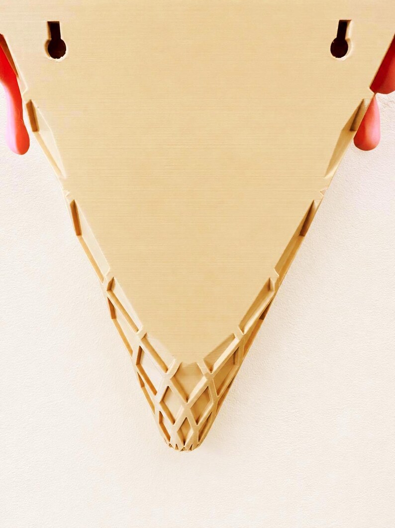 May include: A tan, triangular wall-mounted shelf shaped like an ice cream cone. The surface features a textured, waffle-like grid pattern at the pointed base and two keyhole slots at the top for mounting. Pink, rounded accents protrude from the sides.