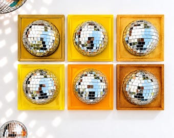 May include: Six square wall panels, each featuring a centered, spherical disco ball made of small mirrored tiles. The frames vary in shades of yellow, gold, and orange. A separate, detached disco ball sits in the lower left corner.