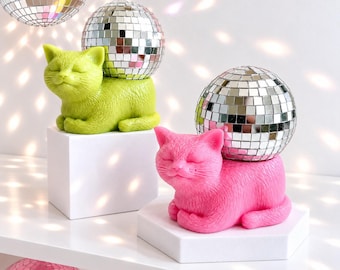 May include: Two cat figurines, one lime green and one bright pink, each resting on a white block. Each cat supports a spherical disco ball made of small mirrored tiles on its back. The cats have closed eyes and a serene expression.