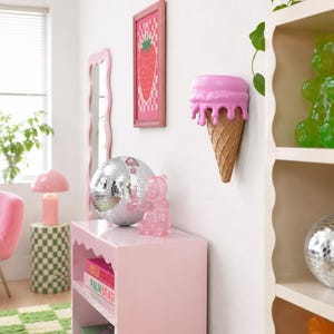 May include: A wall-mounted shelf shaped like a melting pink ice cream cone with a textured tan waffle cone base. The shelf is positioned on a white wall, featuring a smooth, rounded pink top section that appears to drip over the cone structure.