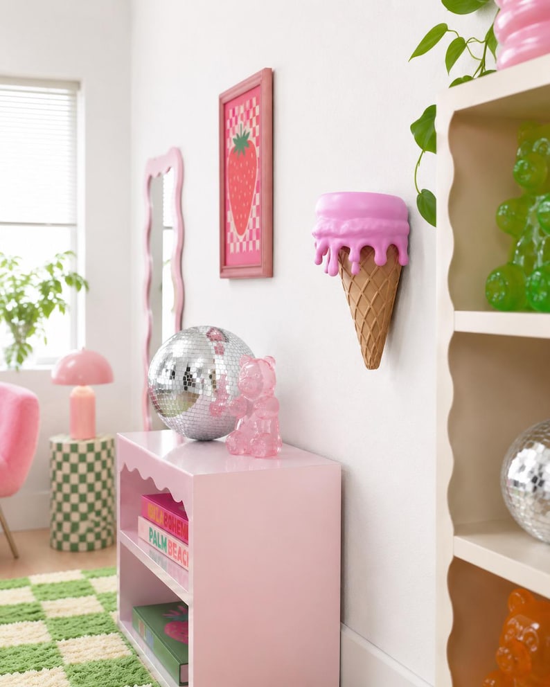 May include: A wall-mounted shelf shaped like a melting pink ice cream cone with a textured tan waffle cone base. The shelf is positioned on a white wall, featuring a smooth, rounded pink top section that appears to drip over the cone structure.