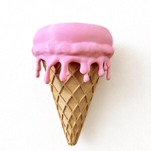May include: A wall-mounted sculpture shaped like an ice cream cone with a tan, diamond-patterned waffle cone base and a large, rounded scoop of pink ice cream on top, featuring thick, realistic drips cascading down the sides of the cone.