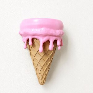 May include: A wall-mounted decorative object shaped like an ice cream cone with a tan, waffle-textured base and a smooth, bright pink scoop on top. The pink scoop features realistic drips cascading down the sides of the cone, creating a dimensional effect.