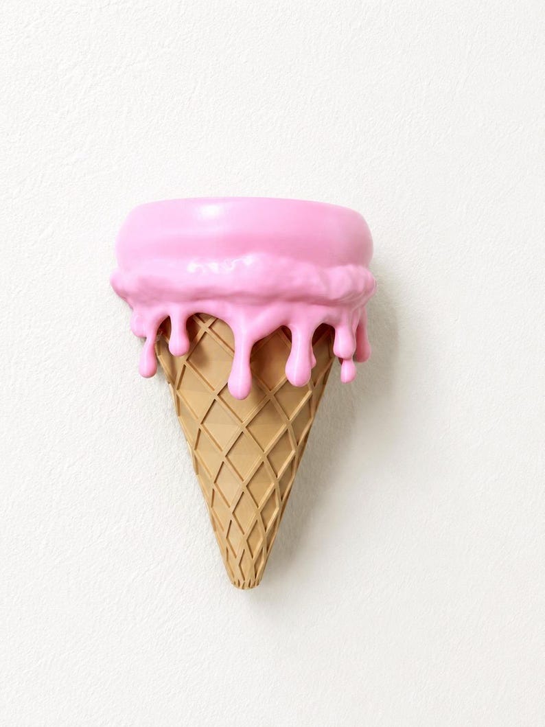 May include: A wall-mounted decorative object shaped like an ice cream cone with a tan, waffle-textured base and a smooth, bright pink scoop on top. The pink scoop features realistic drips cascading down the sides of the cone, creating a dimensional effect.