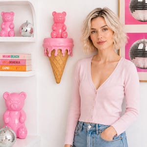 May include: A light pink long-sleeve button-down cardigan paired with light blue denim jeans. The cardigan features a V-neckline and a ribbed knit texture, worn with a simple, clean aesthetic that complements the surrounding decor.
