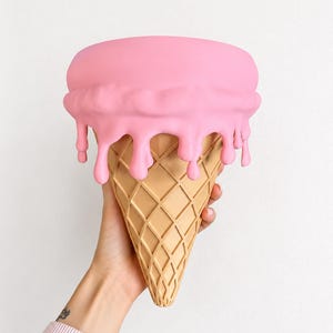 May include: A decorative ice cream cone sculpture featuring a light tan waffle cone base with a textured grid pattern and a smooth, matte pink scoop on top with realistic drips cascading down the sides of the cone against a plain white background.