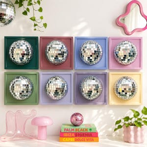 May include: Eight square wall frames in various colors, each containing a silver mirrored disco ball. Below, a stack of three books titled Palm Beach, Ibiza Bohemia, and Capri Dolce Vita sits next to a small pink disco ball and decor.