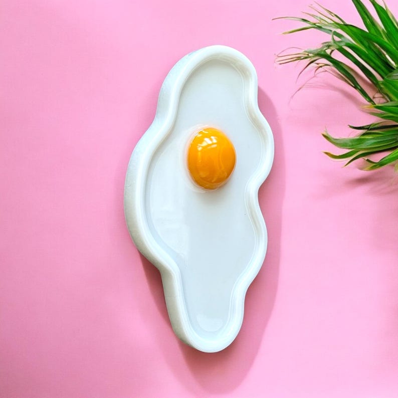 Puede incluir: Una bandeja de cer&aacute;mica blanca con forma de huevo frito y contorno irregular y ondulado. El centro presenta un elemento amarillo esf&eacute;rico, brillante y elevado. La bandeja tiene una superficie lisa y reflectante con una forma org&aacute;nica.