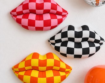 May include: Three wall-mounted lip-shaped sculptures featuring a checkered pattern. One set of lips is pink and red, one is black and white, and one is yellow and orange. Each piece has a three-dimensional, rounded, and plush-like appearance.