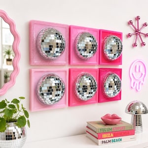 May include: Six square wall art pieces featuring centered silver disco balls mounted on pink frames of varying shades. The arrangement includes a pink wavy mirror, a pink starburst wall decor, a pink neon smiley face, and a disco ball mushroom lamp.