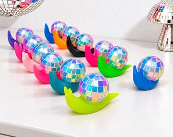 May include: A collection of snail-shaped figurines, each featuring a shell made from a reflective, multi-colored disco ball. The snail bodies are crafted in various solid, vibrant colors including blue, orange, black, pink, green, and yellow.
