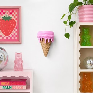 May include: A wall-mounted pink ice cream cone sculpture with a textured waffle pattern. The top section features a smooth, bright pink scoop with realistic drips cascading down the sides, creating a playful and dimensional decorative object.