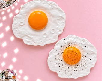 May include: Two ceramic wall decorations shaped like fried eggs. One egg features a smooth, bright orange yolk, while the other has a speckled orange yolk and white surface. Both have irregular, textured white edges resembling cooked egg whites.