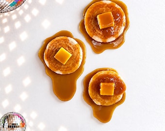 May include: Three round, golden-brown pancakes topped with square pats of yellow butter and drizzled with thick, amber-colored syrup. Two reflective disco balls are positioned in the upper left and lower left corners of the frame.