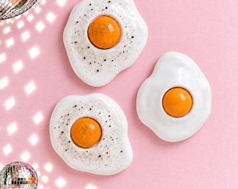 May include: Three ceramic wall decorations shaped like fried eggs. Two eggs feature a white base with a bright orange yolk and black speckles resembling pepper, while the third egg has a smooth white base and a solid orange yolk.