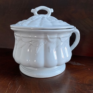 May include: A white ceramic serving dish with a lid. The dish has a handle and a decorative floral pattern. The lid has a loop handle. The dish is on a dark wooden surface.